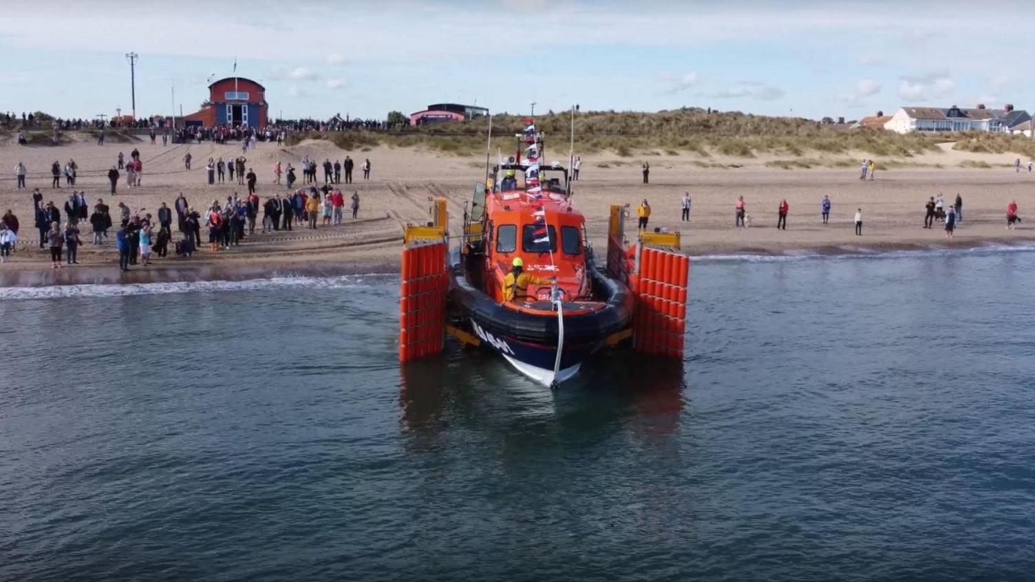 The orange and navy lifeboat enters the sea, with the lifeboat station close by in the background. There are crowds of people on the beach.