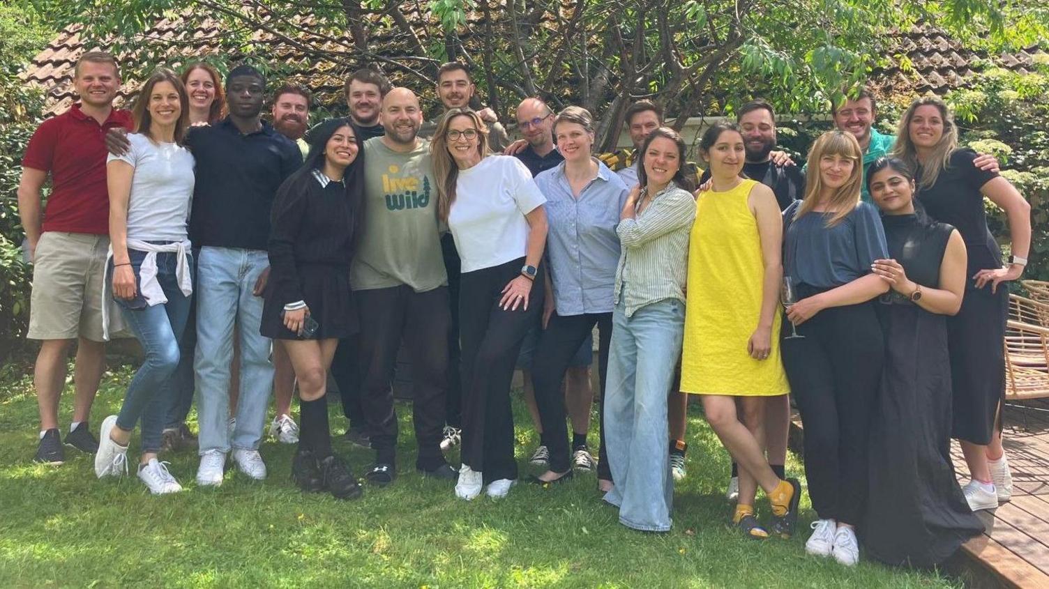 A large group of men and women, about 30 in all, stand on a grassy area in the shade of a tree. They are dressed in summer clothes and it is a sunny day.