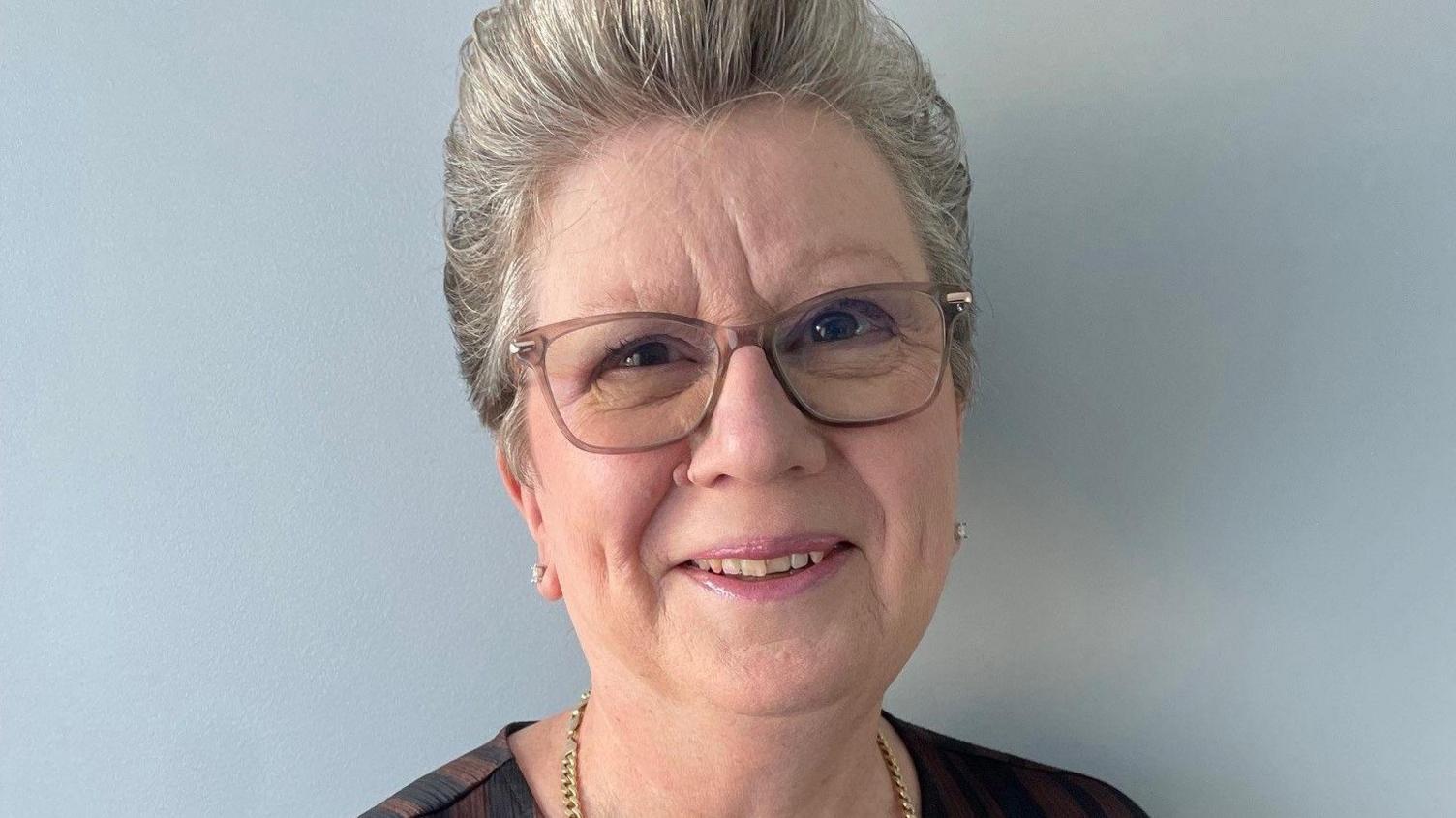 Deena Banister with short grey hair and brown eyes wearing brown glasses and a brown top. She is smiling.