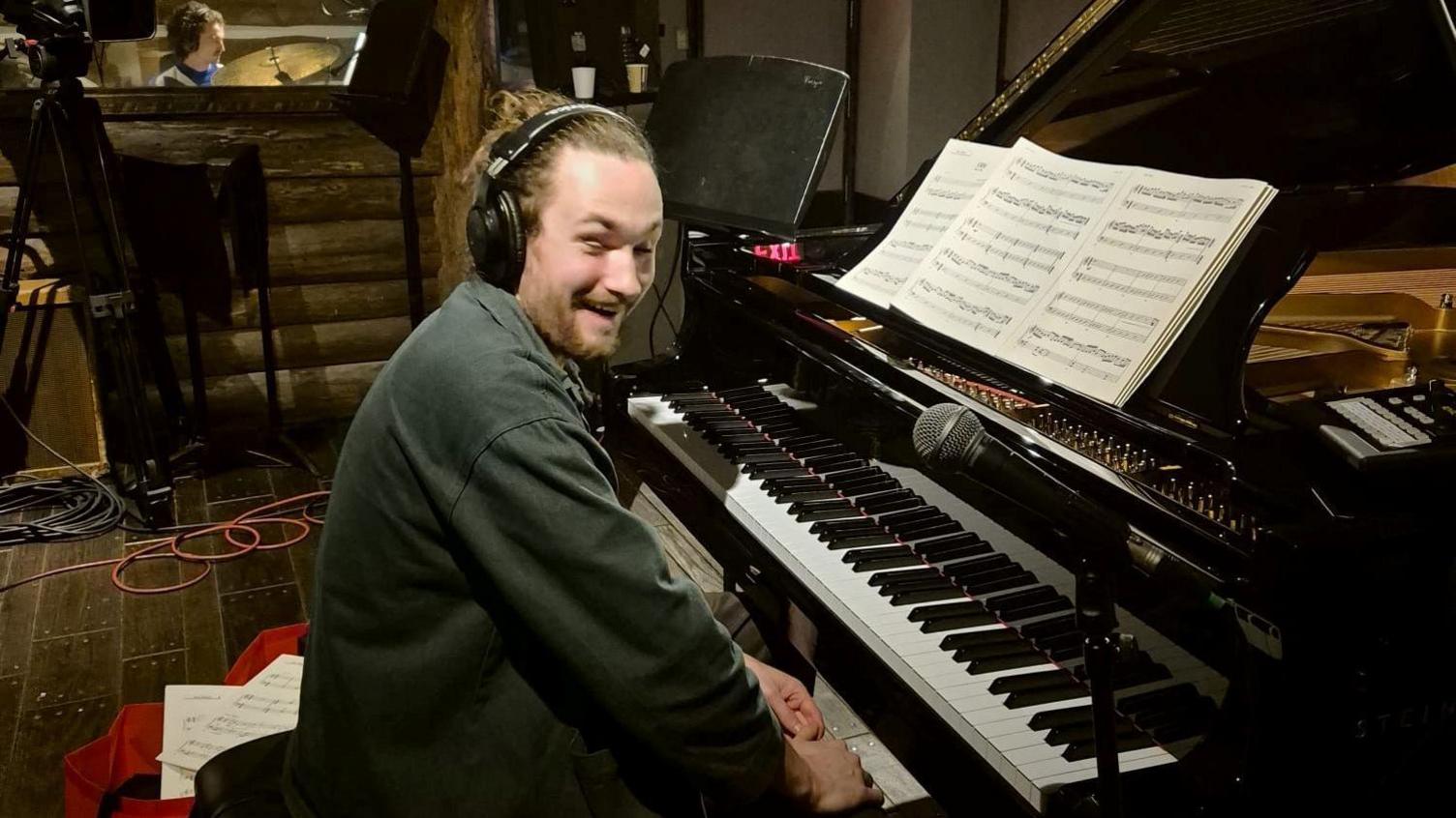A man laughing as he sits at the piano in a recording studio, with sheet music in front of him