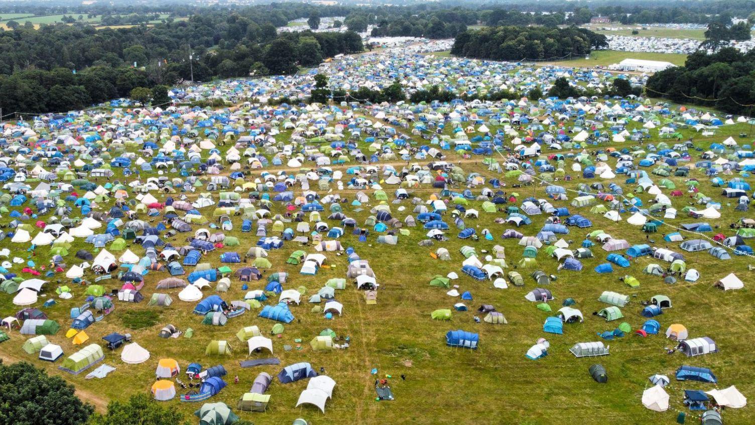 An aerial view of a number of fields showing hundreds of multi-coloured tents with trees in the background.