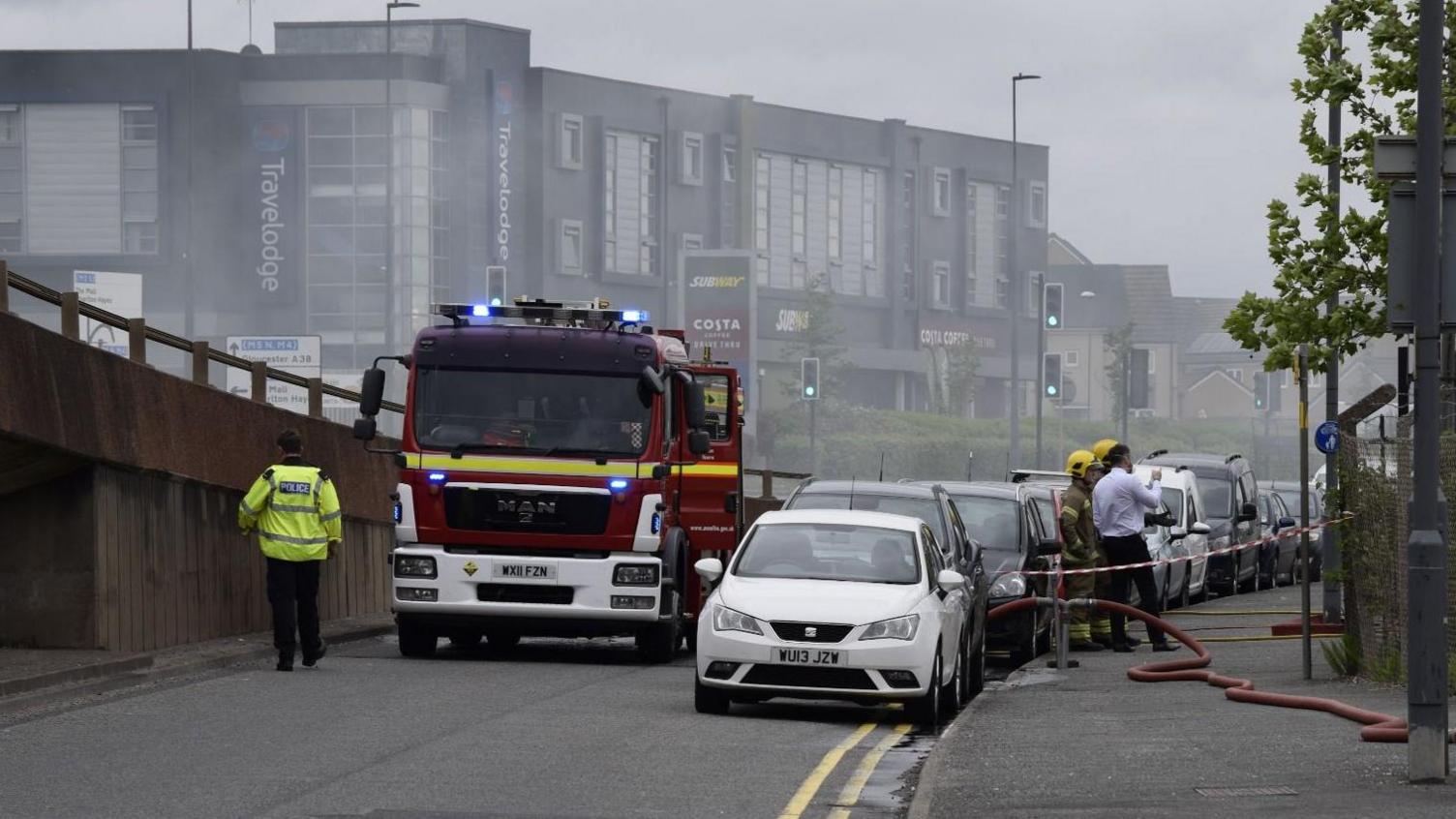 A fire engine and several cars parked along the road. Smoke can be seen in the air