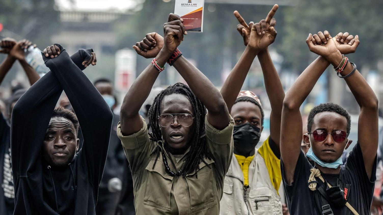 Four male protesters, standing in a row, raise their arms in an "X" symbol.