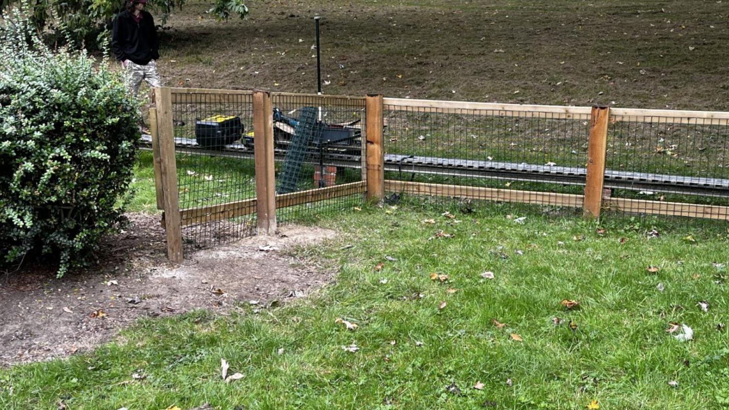 A light railway line running through a park with a green lawn. Alongside the line is a set of wooden fencing built to roughly waist height, with chicken wire between the top fence and the ground.