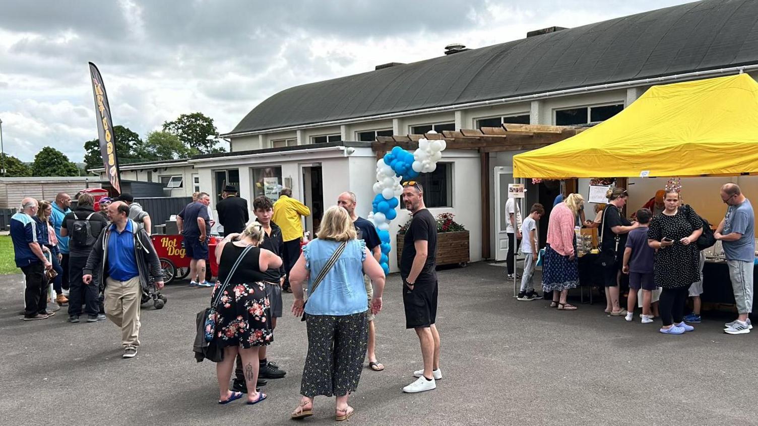 Many people gathered outside a building in Westbury. A yellow canopy is up above a table on the right and a hot dog stand can be seen to the left. People are gathered around, at the table and having conversations.