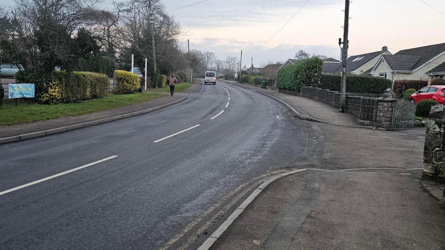 A residential road going through the village of Tutshill. There are houses on either side and a pedestrian is walking in the distance.