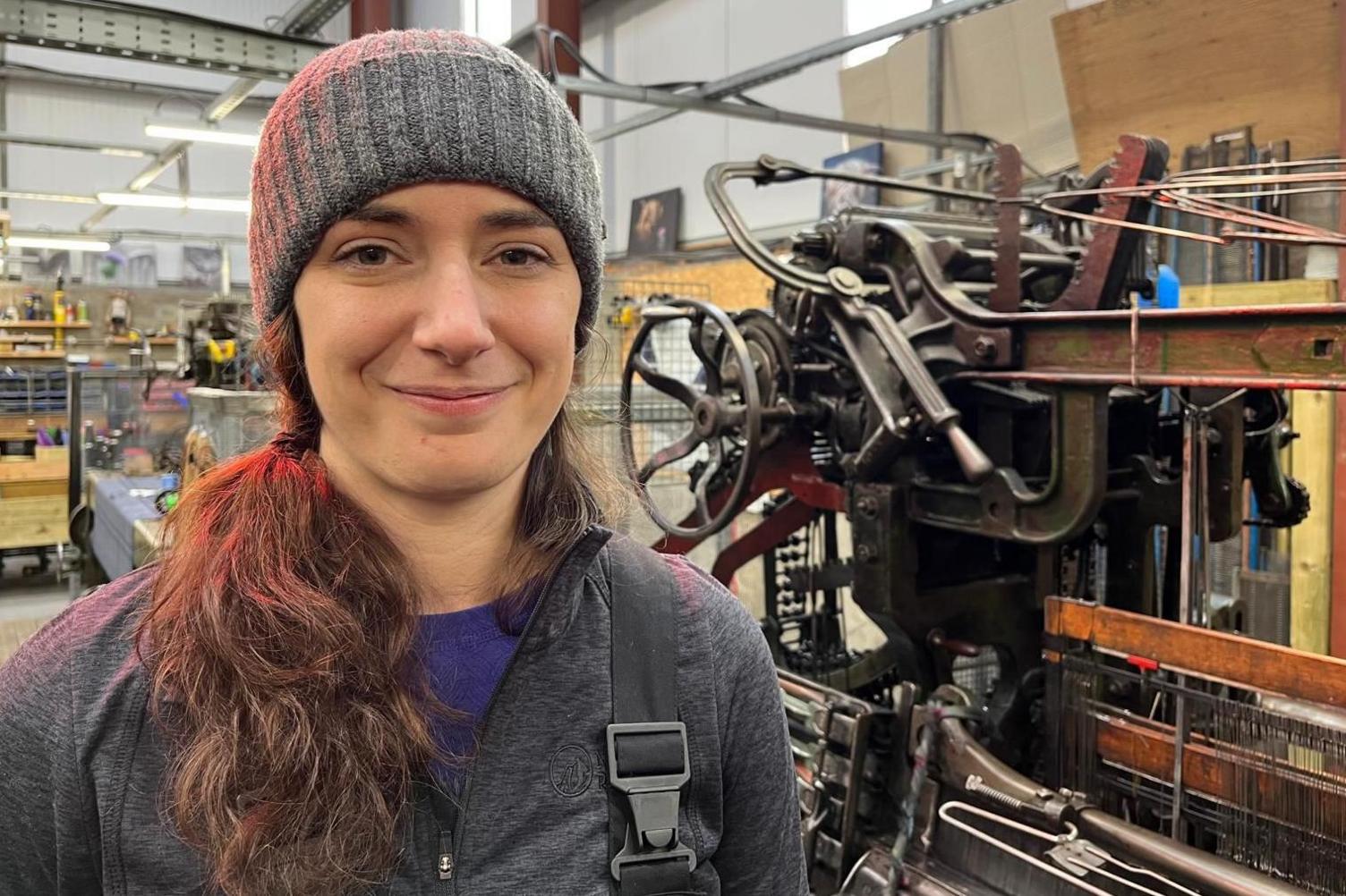 Robyn is standing next to a loom at Prickly Thistle's mill. She is wearing a woolly hat and is smiling.