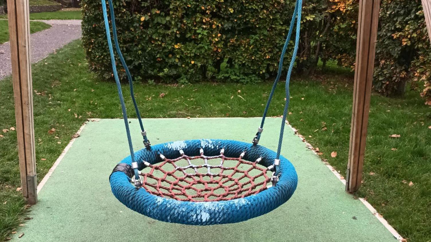 A blue and red basket swing with light blue paint splattered across it. Underneath the swing is green tarmac and the swing is held up with wooden beams. Around the tarmac is grass and a hedge behind it