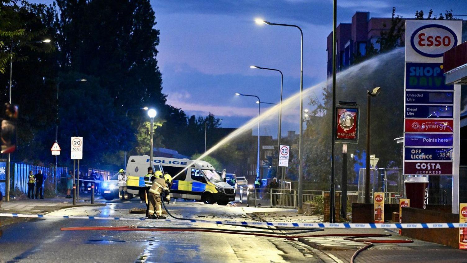 Two firefighters, in protective clothing, spray water on to the fire at the derelict pub. A police cordon and van are in the background. The fire happened overnight and the sky is starting to get brighter as the sun rises.