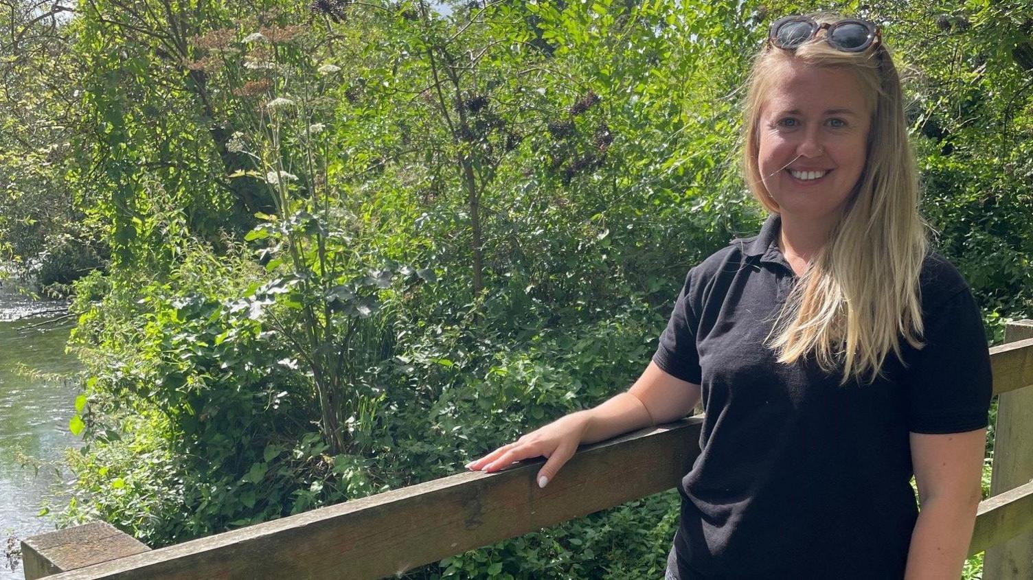 Sarah Perry, standing on a bridge across the River Lea, smiling at the camera. She is wearing a dark polo shirt and sunglasses on her head.