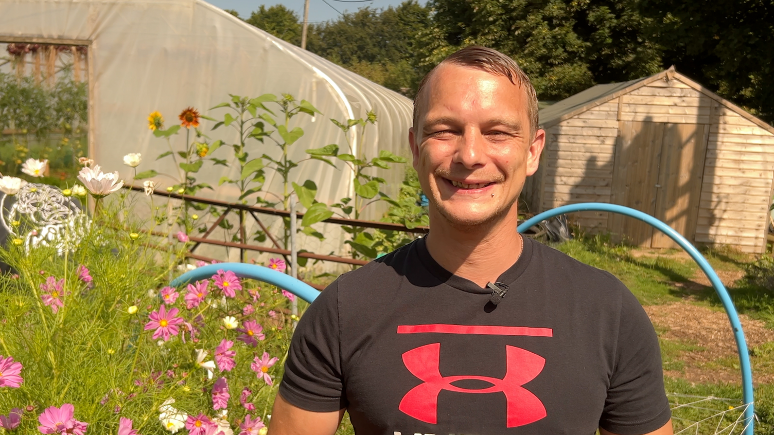 A man smiles into camera. He's wearing a black top with a red logo. Behind him is a shed and a polytunnel.
