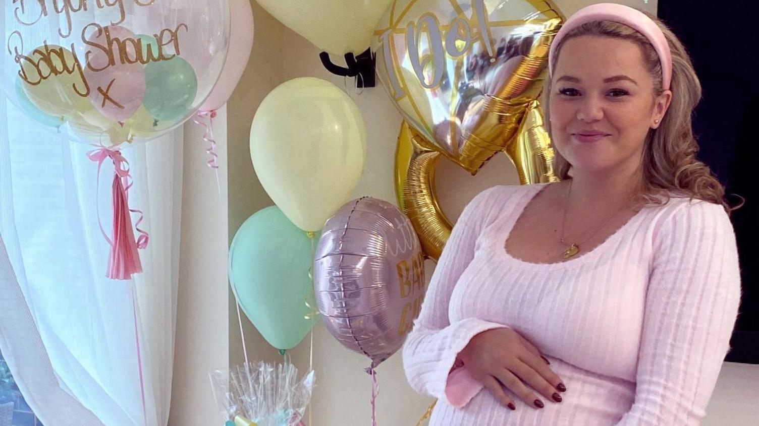 Bryony Russo wearing pink lounge clothes and a headband. She is smiling and holding her baby bump at a baby shower, with lots of balloons behind her.