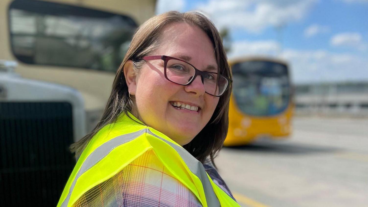 A middle aged woman smiles as she watched a yellow single decker bus