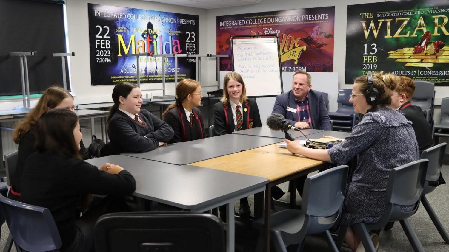 A number of school pupils wearing black blazers with red trimming, black jumpers, and red and green ties sit around a table in a classroom. A man sits at the head of the table in a pink and navy check pattern shirt and grey blazer. A woman wearing headphones sites holding a microphone pointed to the middle of the table