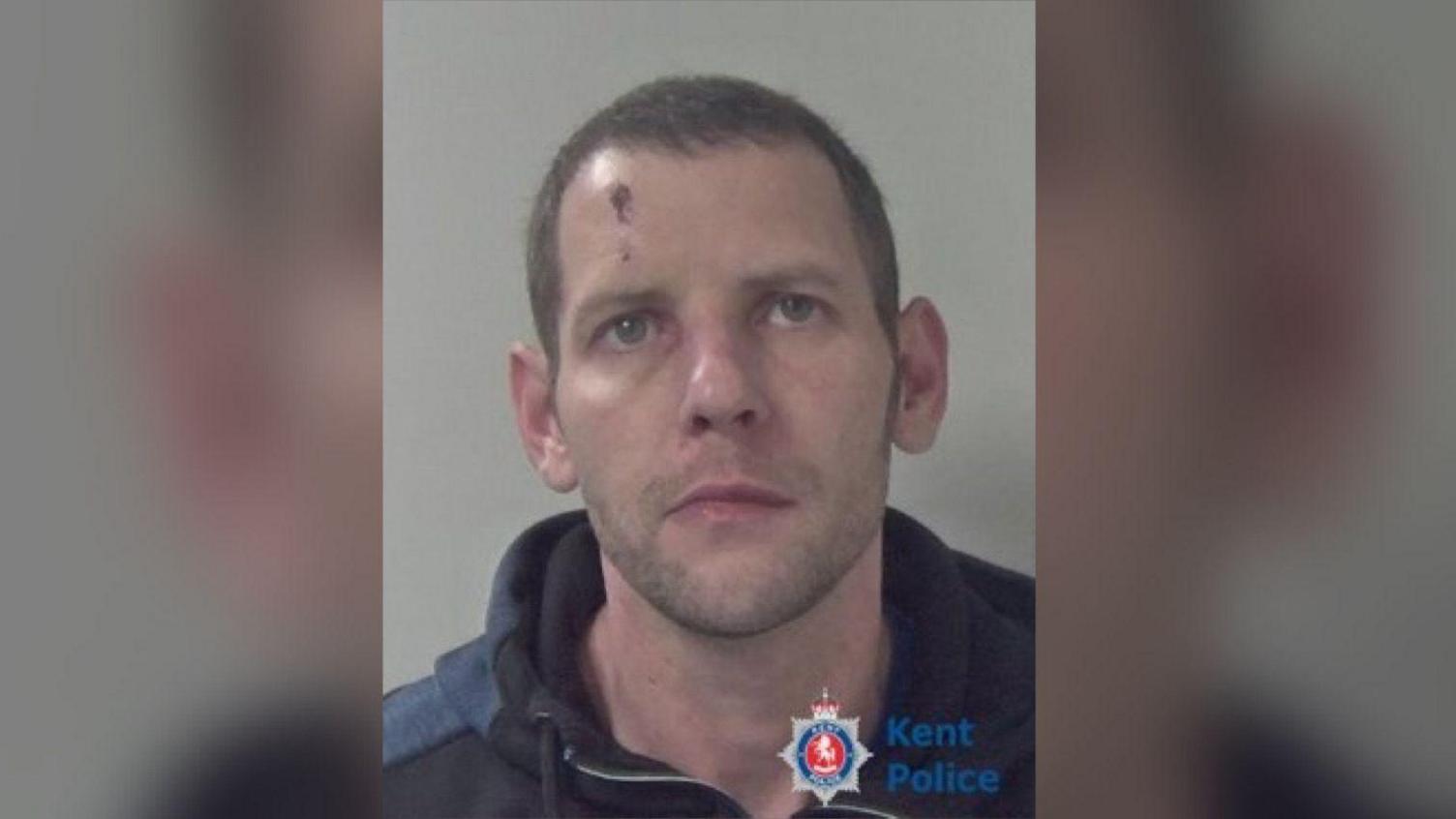 A mugshot of Ajay Porter - a man with a scar on the right of his forehead, short brown hair and stubble. He is wearing a black and blue hoodie.