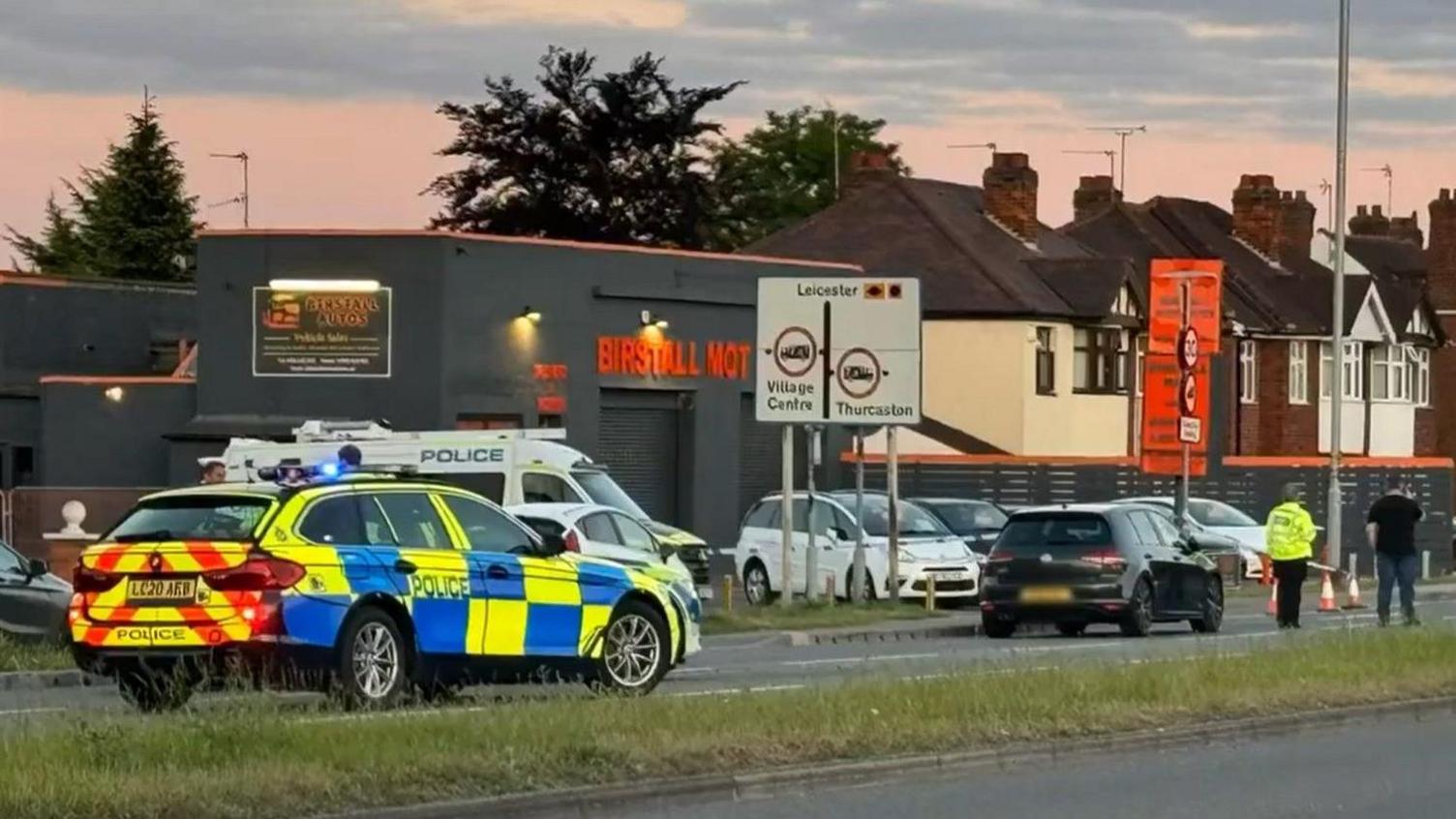 A police car stopped on the A6 in Birstall, outside a garage. A police officer and plain-clothes man stand facing away from the camera, looking at the road