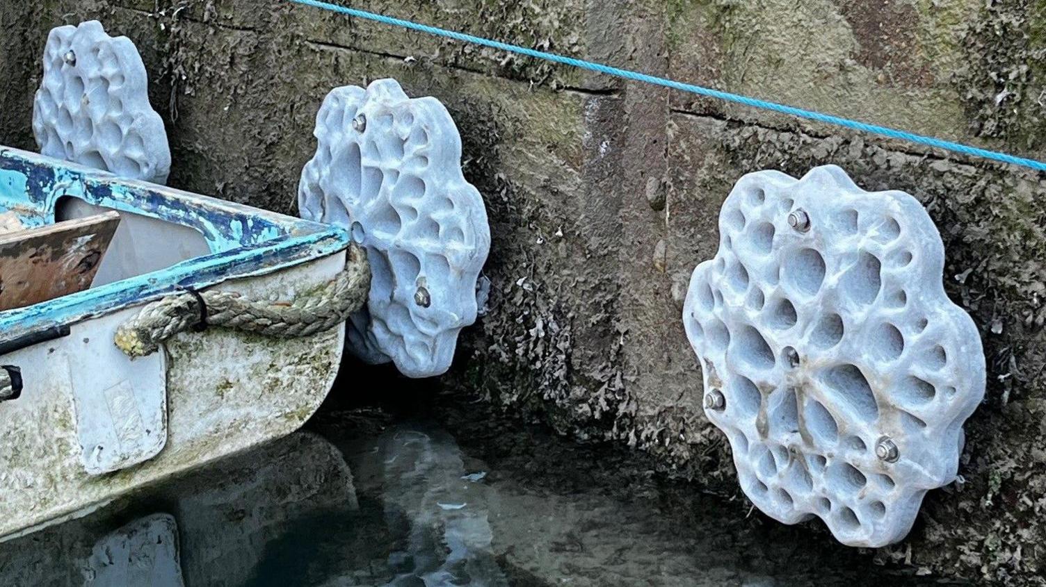 Three sea tiles attached to the wall of a marina just above the water and next to a rowing boat floating in the water.