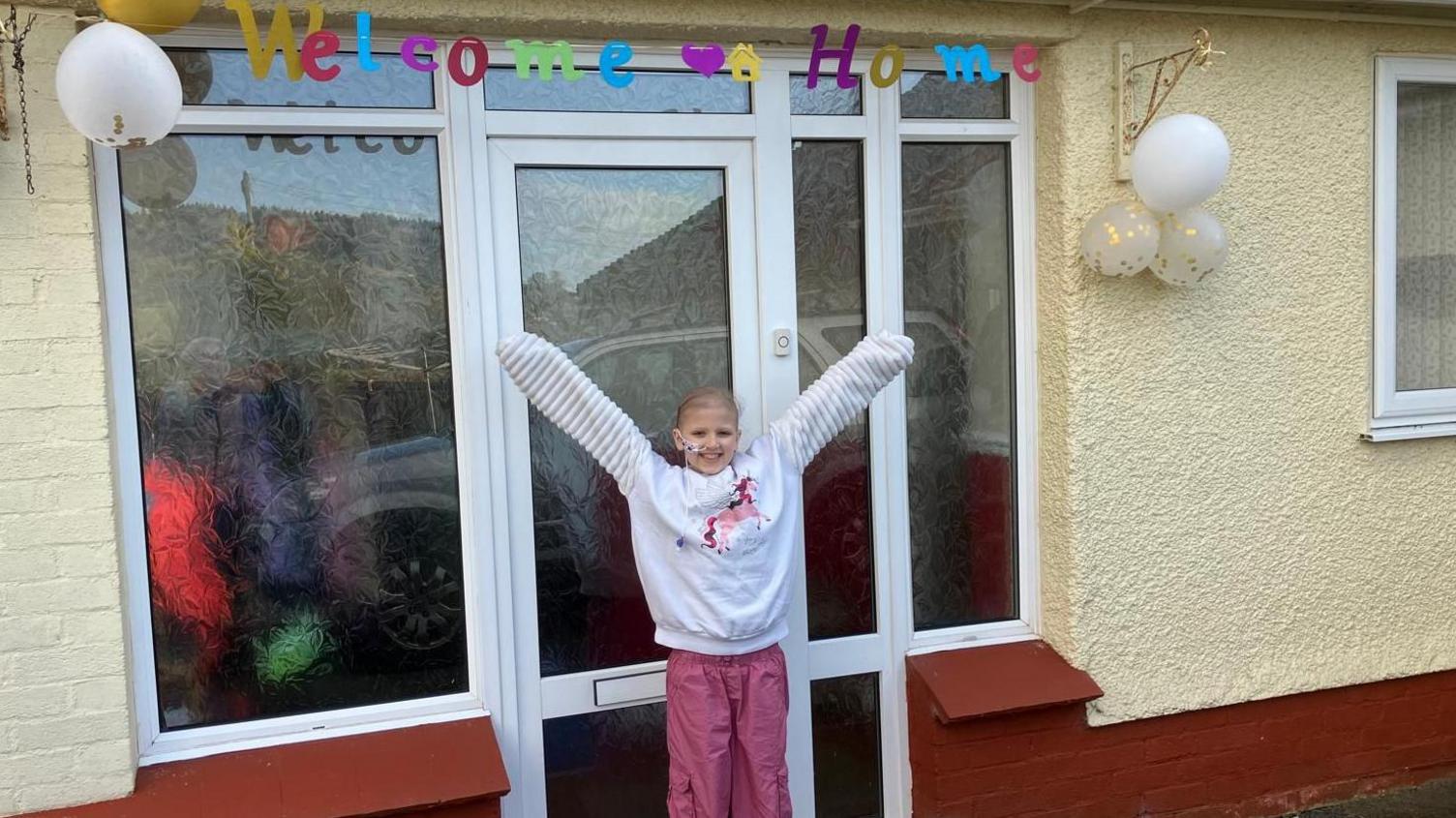 Kathryn yn sefyll o flaen drws ei chartref. Mae'n gwenu ar y camera ac mae ei dwylo hi yn yr awr. Mae'n gwisgo siwper wen a throwsus pinc. Mae yna arwydd 'Welcome Home' uwch ei phen.