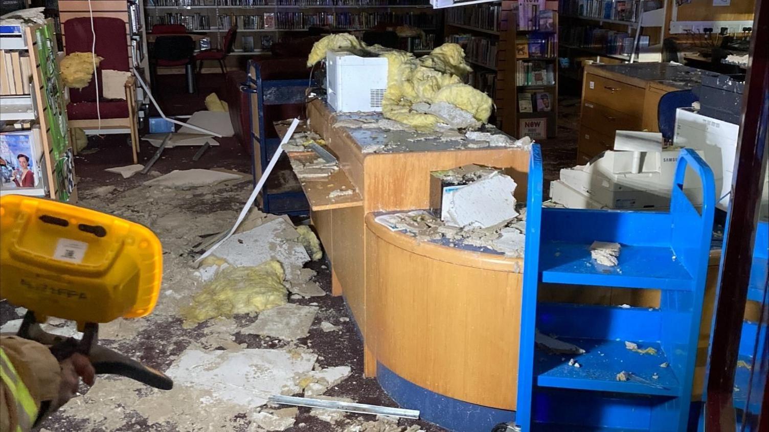 Damaged items are scattered across the library floor and on a wooden countertop. In the background are shelves filled with books.