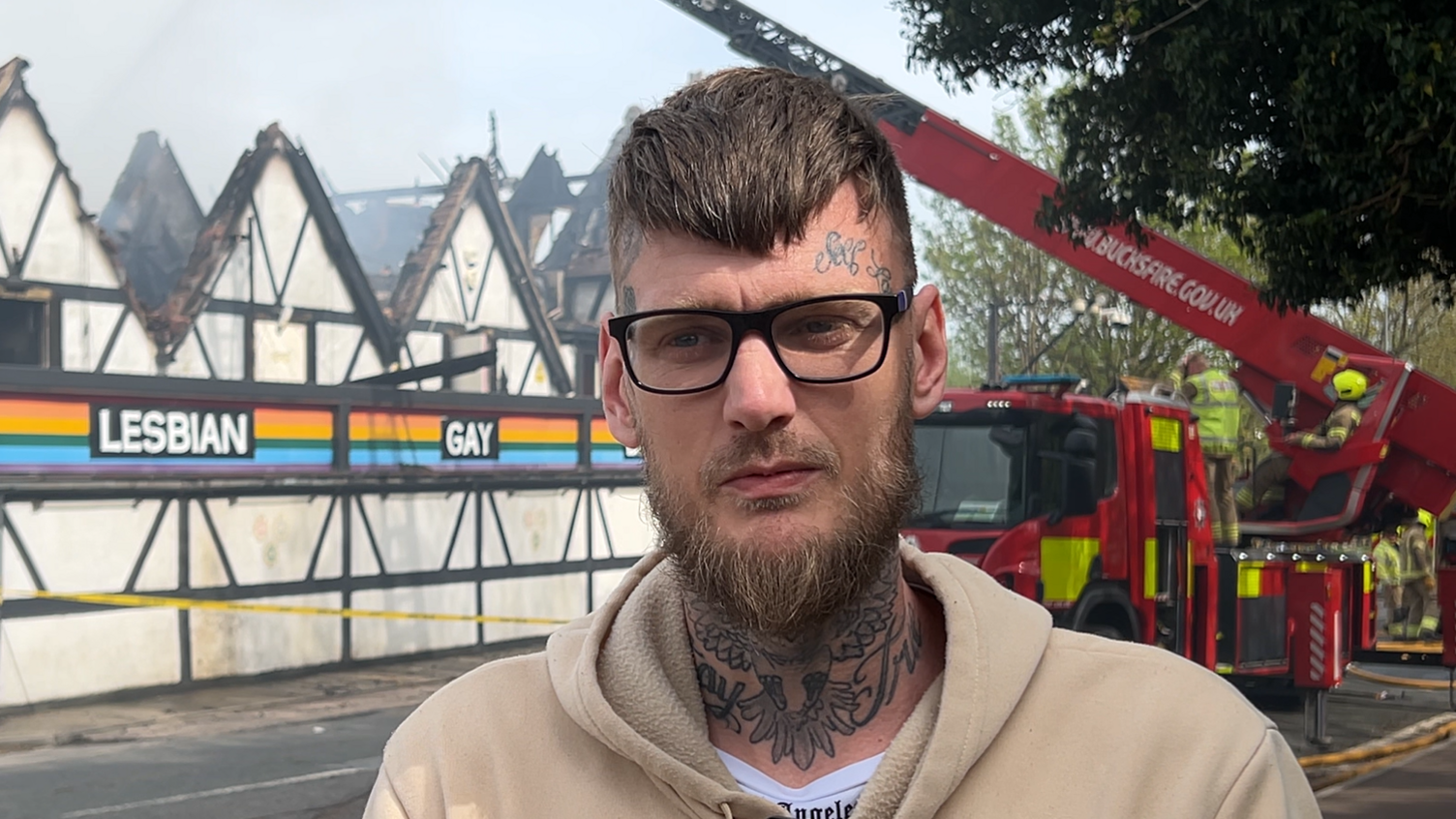 A man with dark hair that rests over his forehead and a thick beard looks slightly to the right of the camera. He wears glasses and a cream hoodie. Behind him is a nightclub which the roof of has been burnt in a fire. A fire engine can be seen behind him with an aerial ladder and firefighters in uniform.