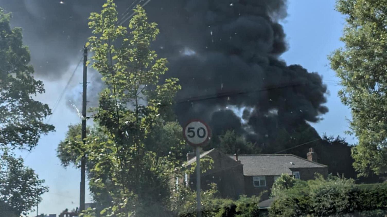 A two-storey residential property in the foreground with black smoke billowing up behind it. The area is surrounded by trees and there is a circular 50 speed limit road sign in front.