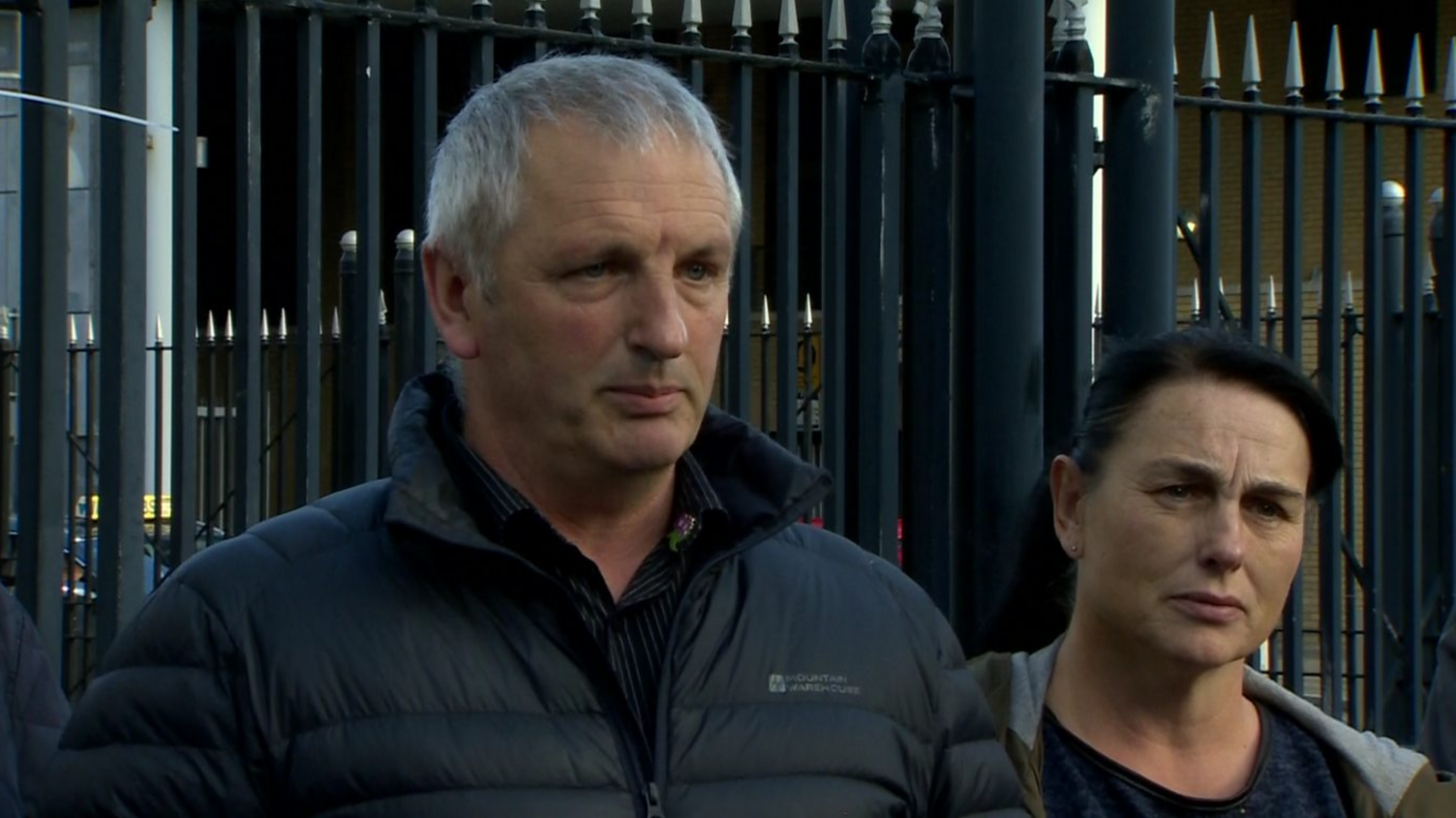 Michael McConville is standing looking slightly off camera with his lips pursed. He is wearing a black coat and black shirt. To his left a woman stands looking on. Behind them are black railings.