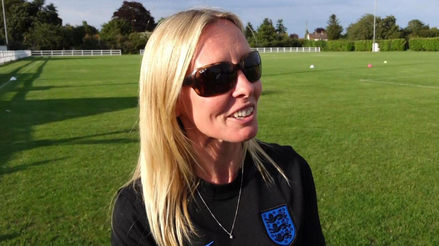 A blonde woman wearing sunglasses and a football shirt gives a television interview with a football pitch in the background.