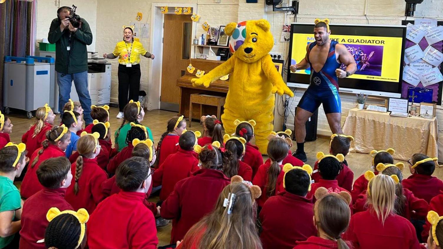 A giant costumed Pudsey bear and TV personality Gladiator Steel in front of around 20 young children with their backs to camera wearing red jumpers and Pudsey ears