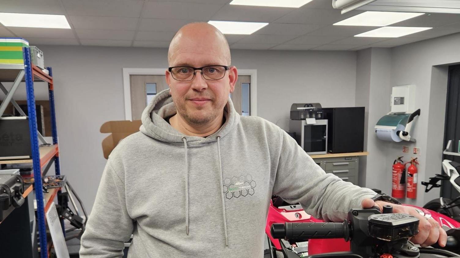 Dan Rack from Drury Precision Engineering stands with his hand on the handlebar of a red motorbike. He is in an office and wearing a grey hoodie with a bald head and black rimmed glasses.