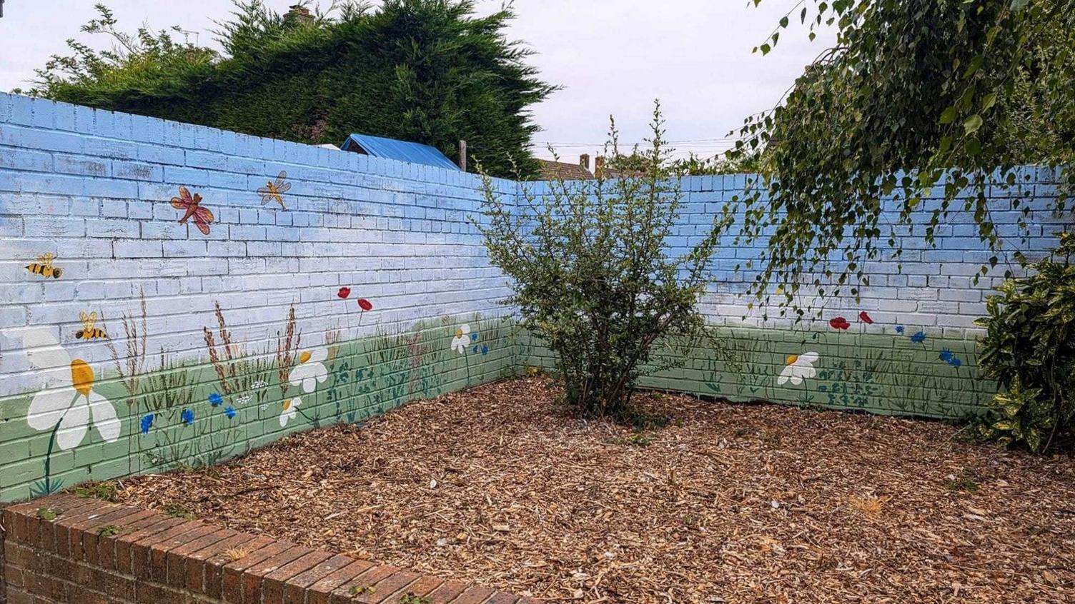 The light blue mural which has been painted onto a brick wall shows bees and dragonflies flying above grass and flowers