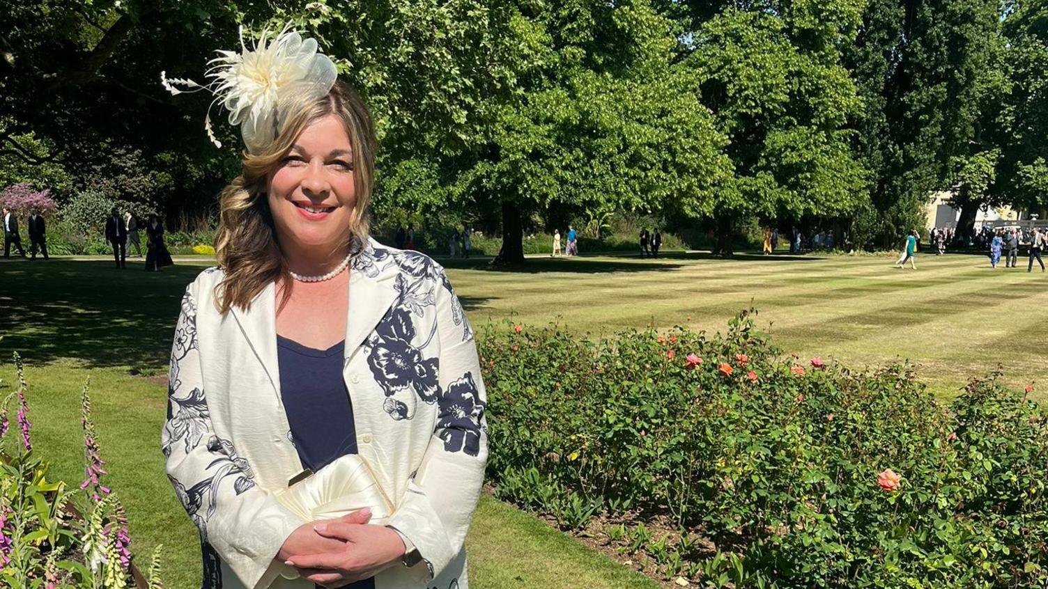 A lady is smiling at the camera. She is smartly dressed in a cream blazer with navy flowering detailing, and also has a matching cream fascinator on her head. She is stood in front of lush greenery of trees, hedges and a well-manicured lawn.