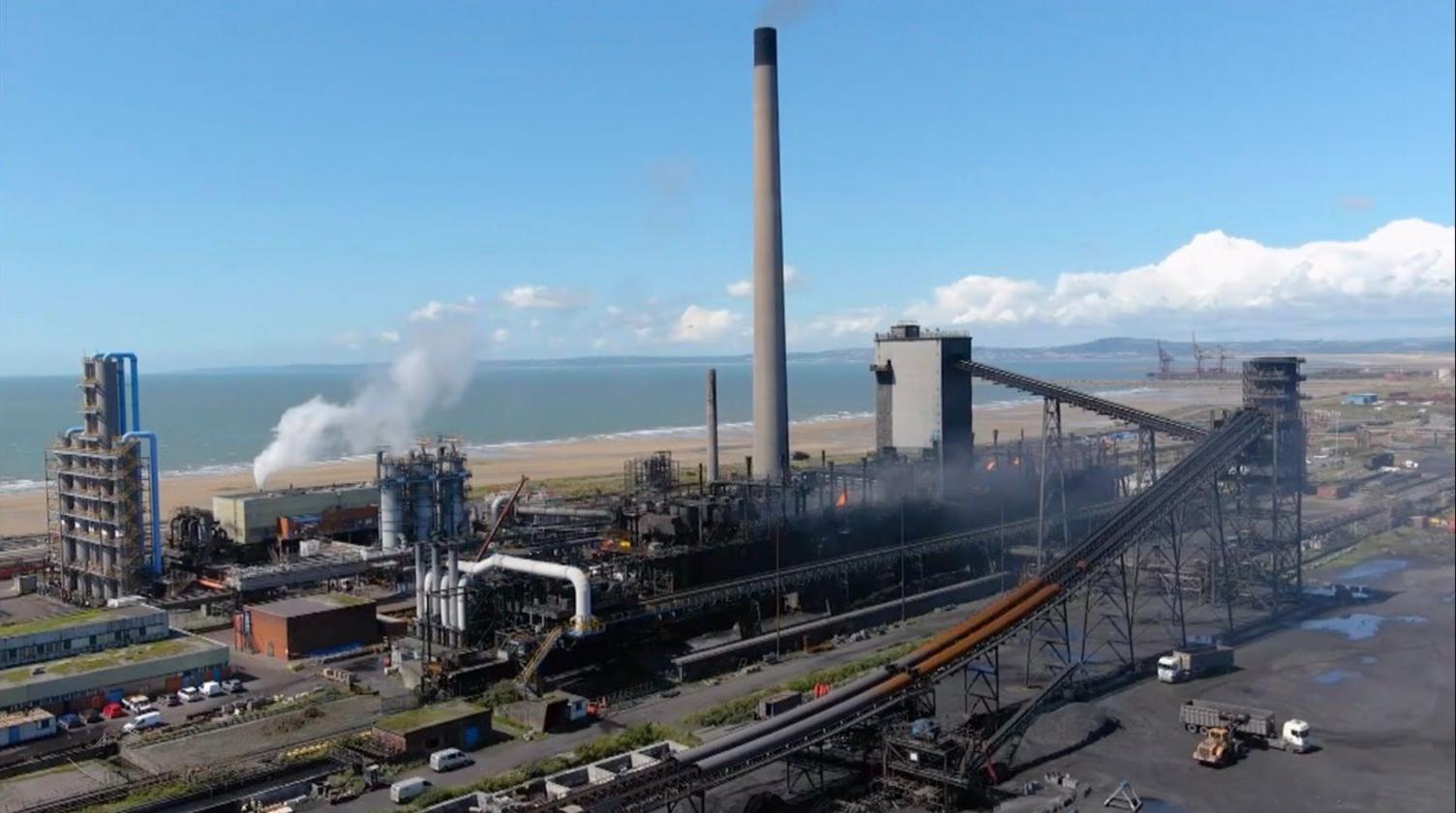 An industrial view which includes extensive large pipework across part of the Port Talbot steelworks site. There is some white smoke coming from various chimneys and flames. There's also a very tall chimney in the centre of the image. In the background there's a beach, the sea along and a blue sky and some clouds.