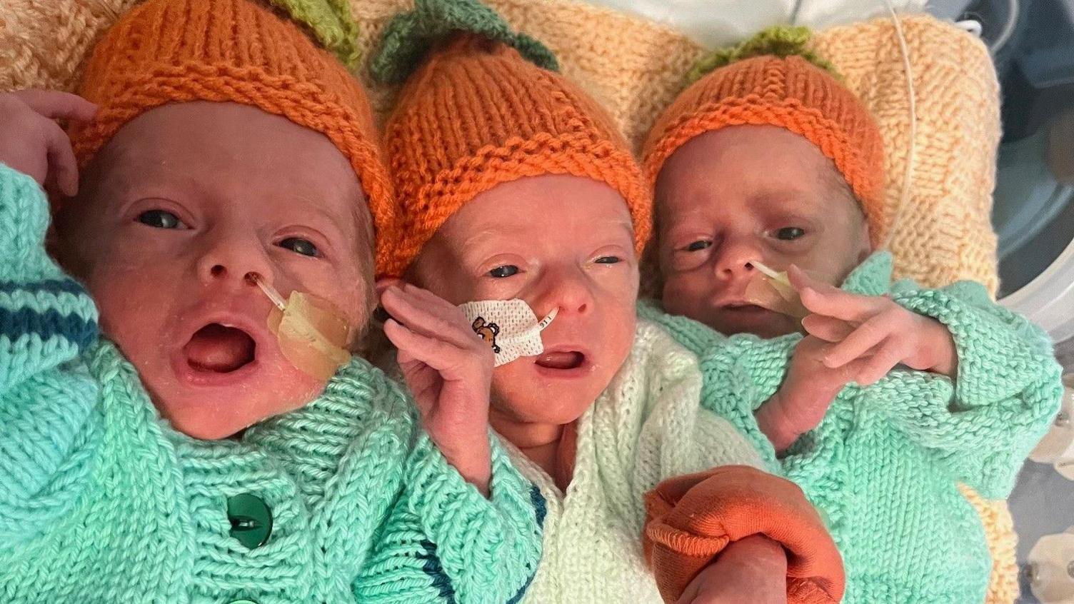 The trio of triplets lie together on a beige-coloured blanket. They are wearing matching green cardigans and knitted orange hats. All three are lying on their backs and have their eyes and mouths open