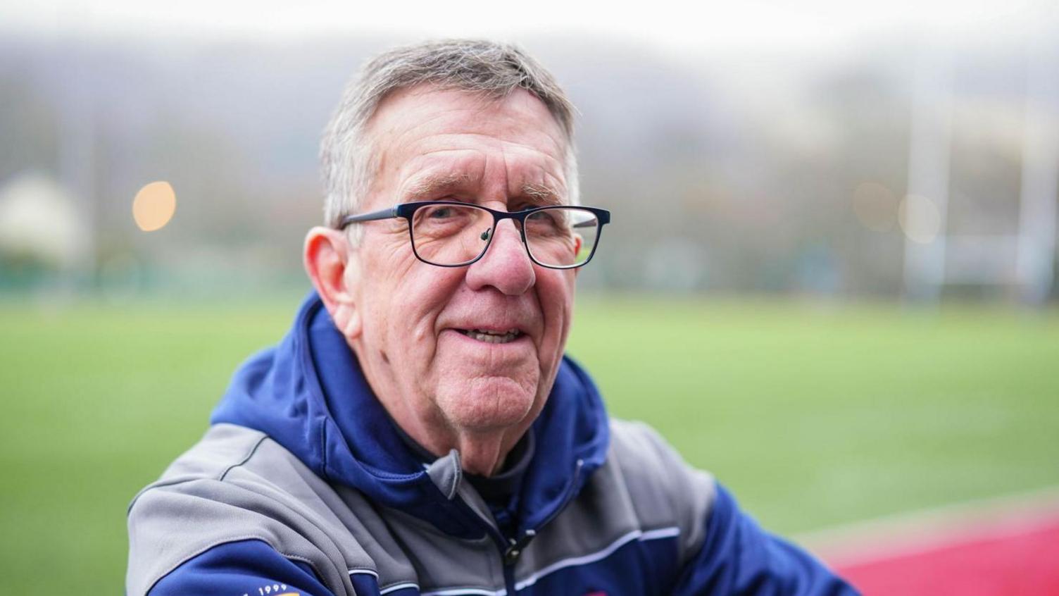 Colin Davies looking at the camera with the rugby pitch behind him. He has grey short hair and is wearing glasses and a blue and grey hoodie. 