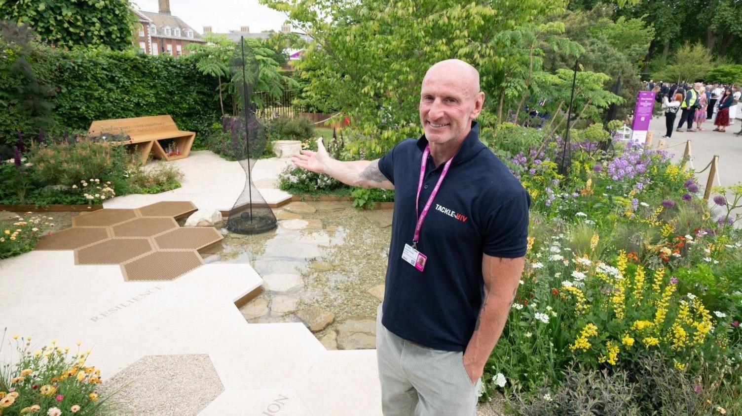 Gareth Thomas smiling in the garden. He is wearing a Tackle HIV T-shirt and has his arm out signalling towards the gardens beauty.