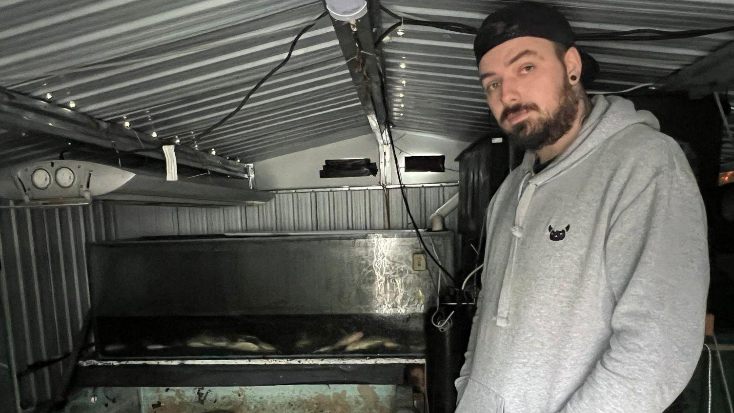 Craig Anderson, who is wearing a grey hoodie and black snapback cap, stands inside a shed next a counter of dead fish above a tank.