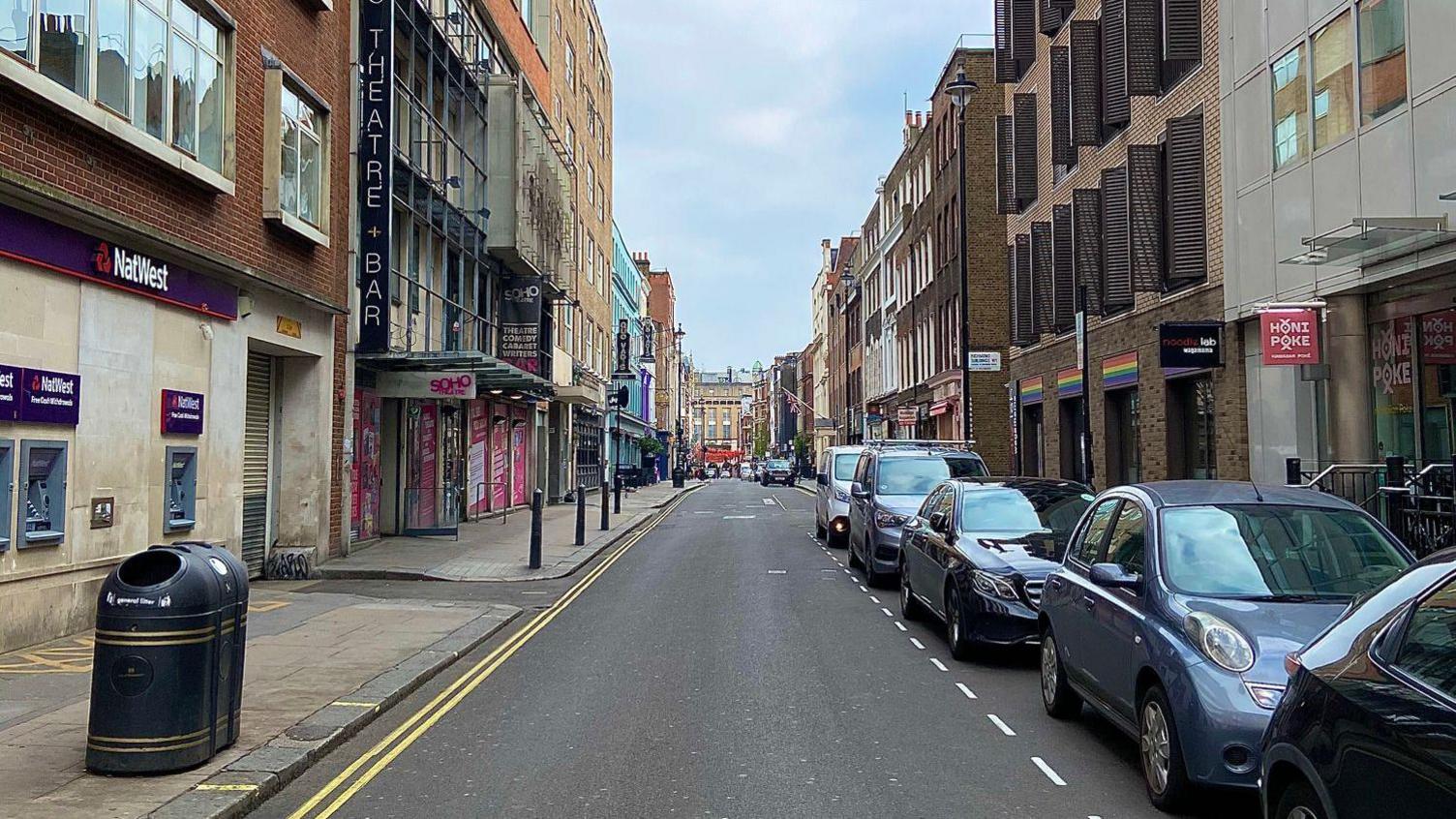 Image of Dean Street in 2020, with a row of parked cars on the right, and black bins and signs for a theatre and bank on the left