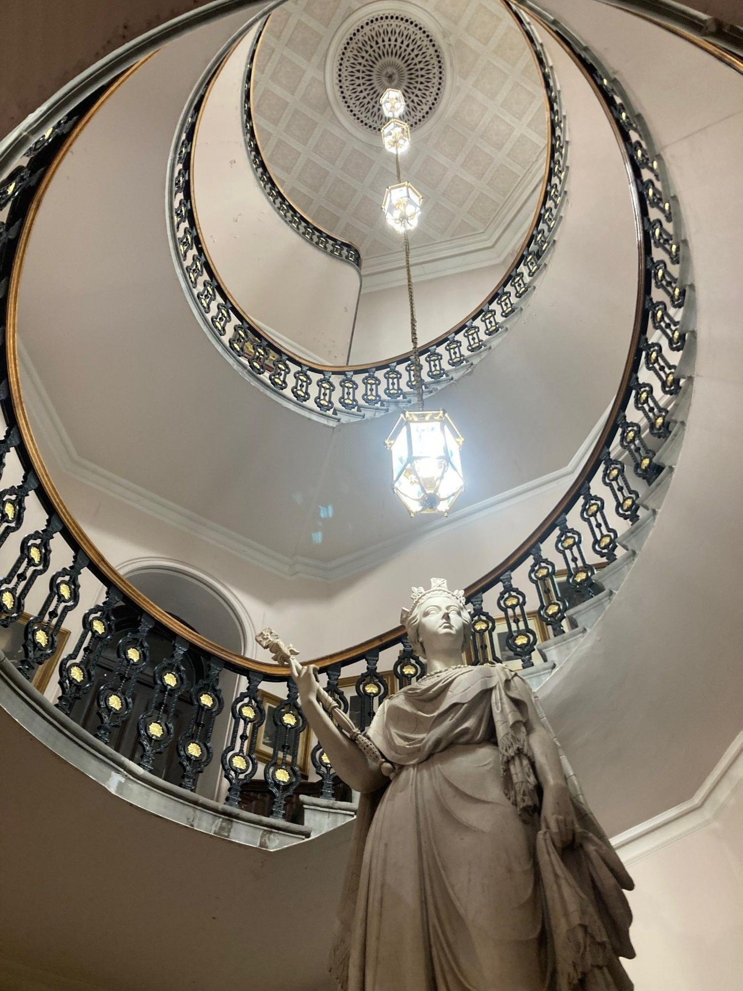 A marble statue of Queen Victoria, photographed from below, with a winding staircase descending up and an ornate light hanging from the ceiling