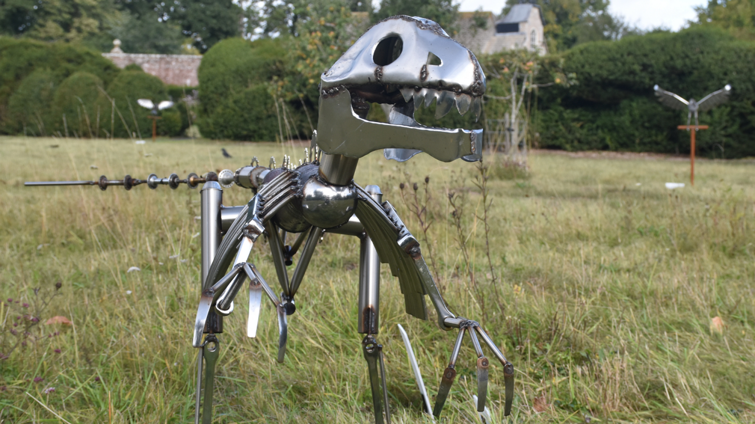 A metal sculpture made to look like a dinosaur standing in a meadow-type area. Behind it other metal sculptures can be seen in the distance including a bird of prey. The meadow is ringed with a hedgerow