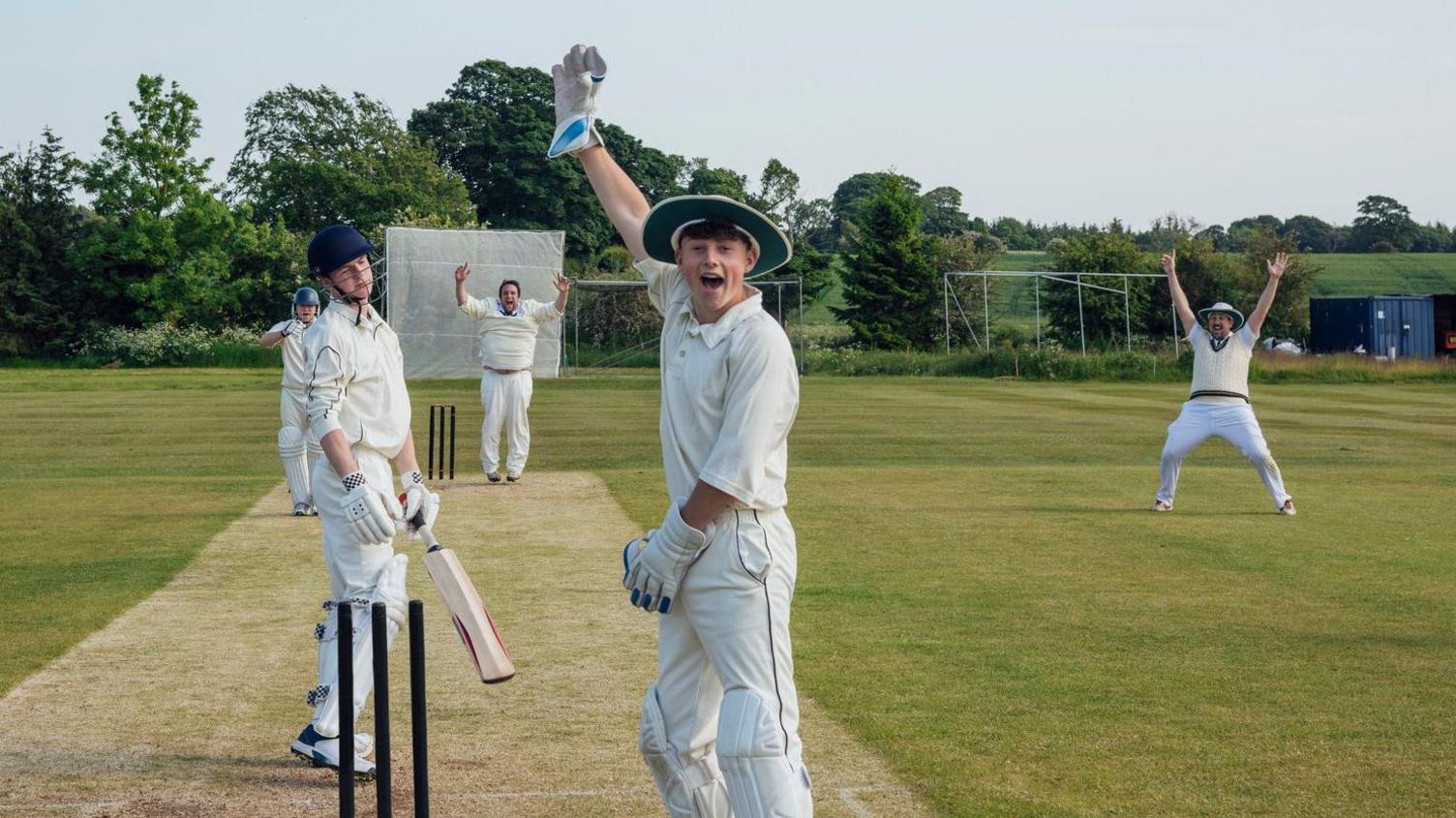 A group of teenage boys play cricket on a green. They are wearing cricket whites and holding their hands up in the air.