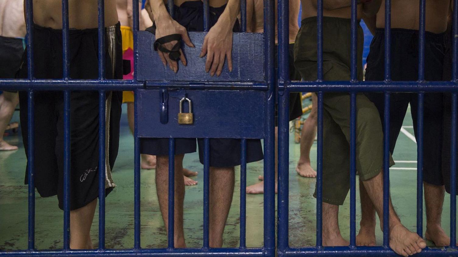 Foreign detainees stand behind bars at an immigration detention centre in Bangkok.They are wearing dark-coloured shorts and pants, with some of their bare torsos and legs visible. One man's hands are also visble above the lock on the cell door.  