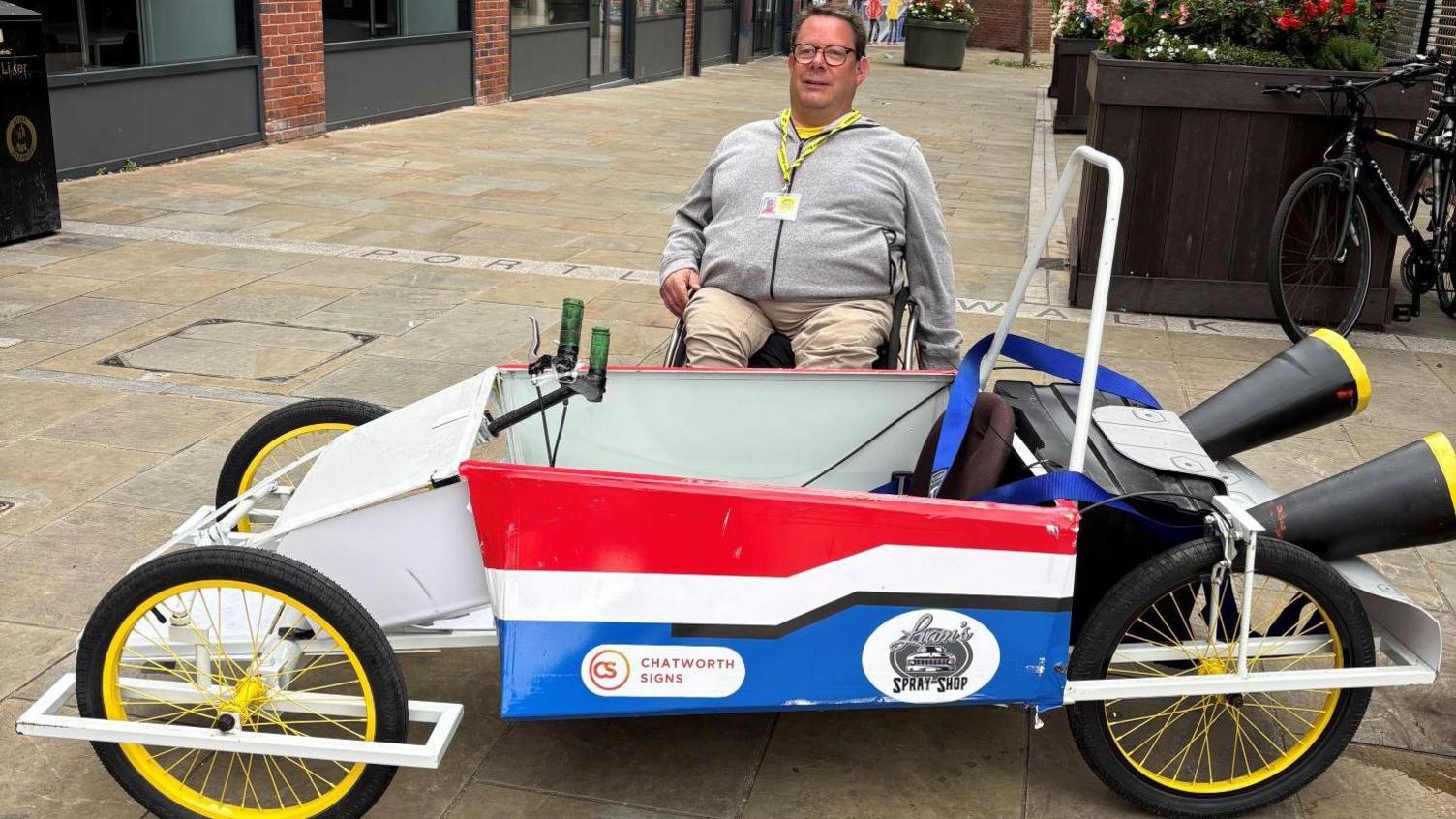 The cart is painted red, white and blue and is made up to look like it has large exhaust pipes at the back. A man behind it is smiling at the camera.