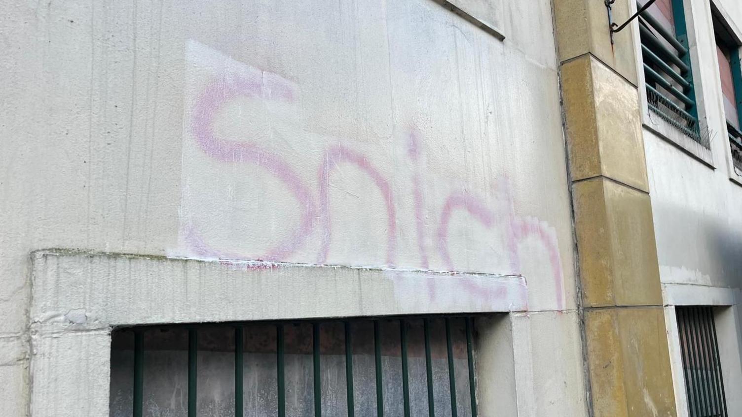 A grey wall with graffiti spelling the word "snich" in pink lettering. There are several cut-outs in the wall with black bars over them.