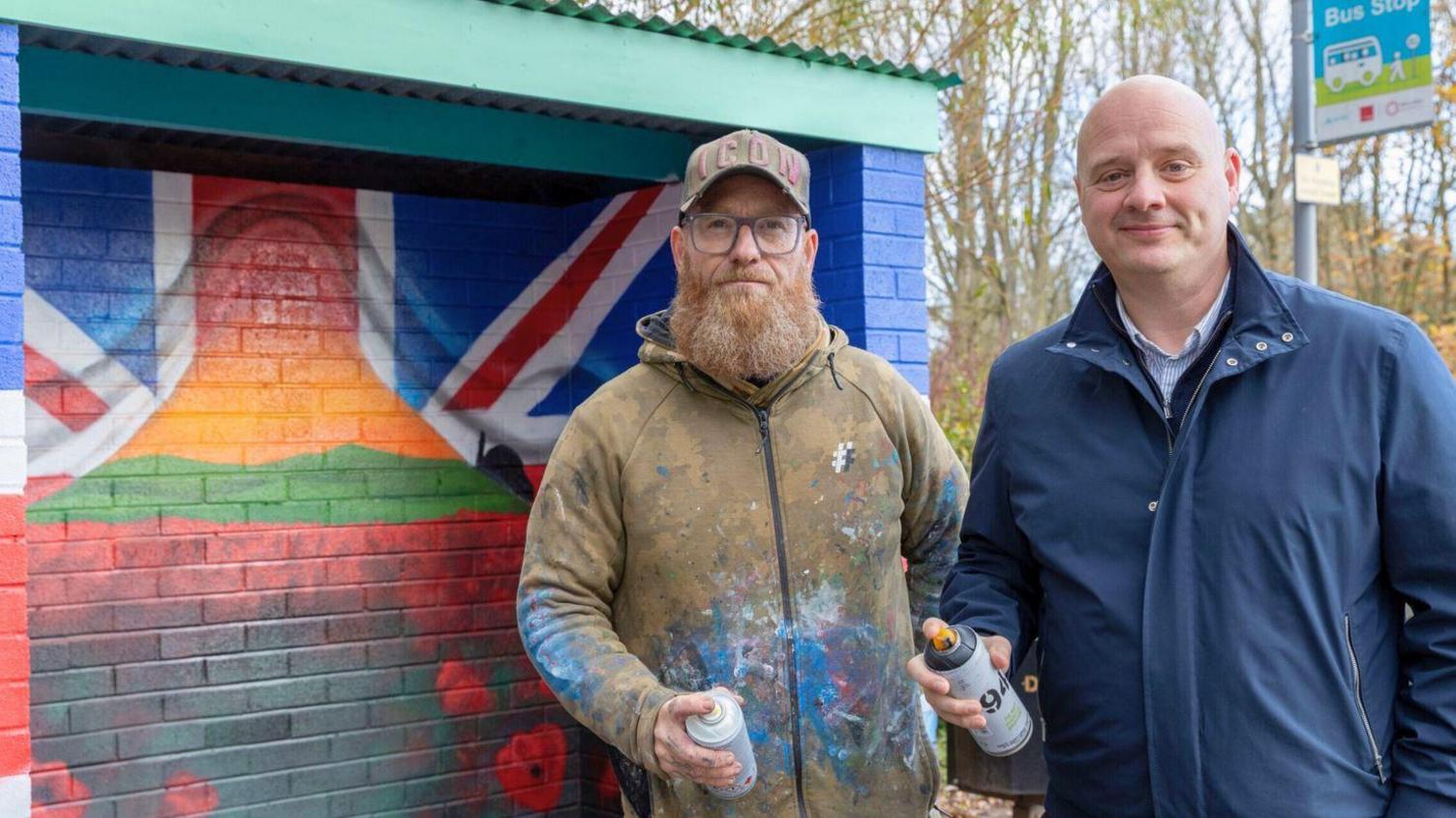 Two men holding white aerosol cans in front of a painted bus shelter. One of them has a brown, paint-covered coat, cap and brown beard and the other has a bald head and blue jacket.