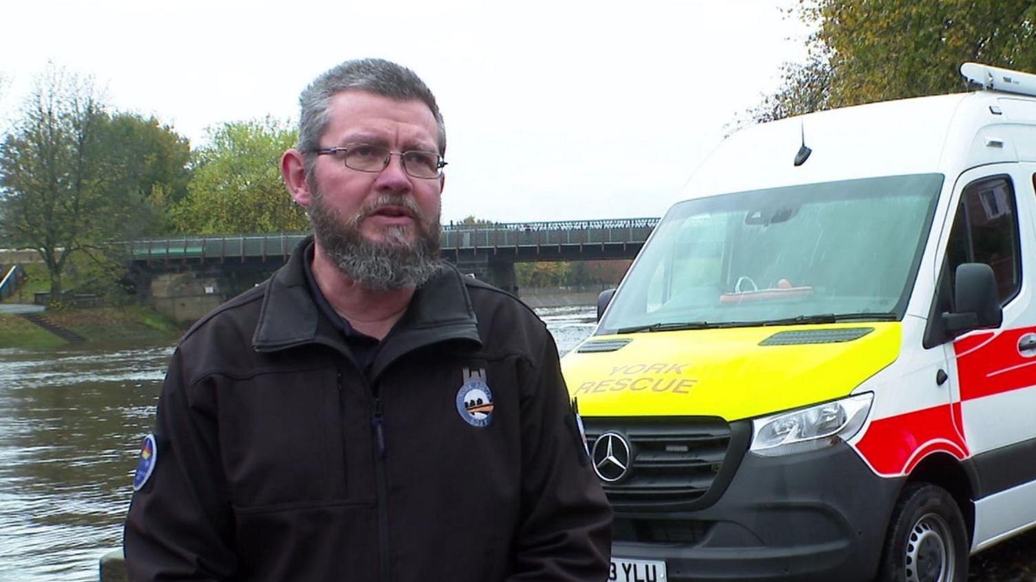 David Wilson stands in front of the wide River Ouse. He wears a dark jacket with York Rescue Boat logos. To his left is white York Rescue van, with a yellow bonnet and red and white pattern down the side. In the background are trees and a bridge.