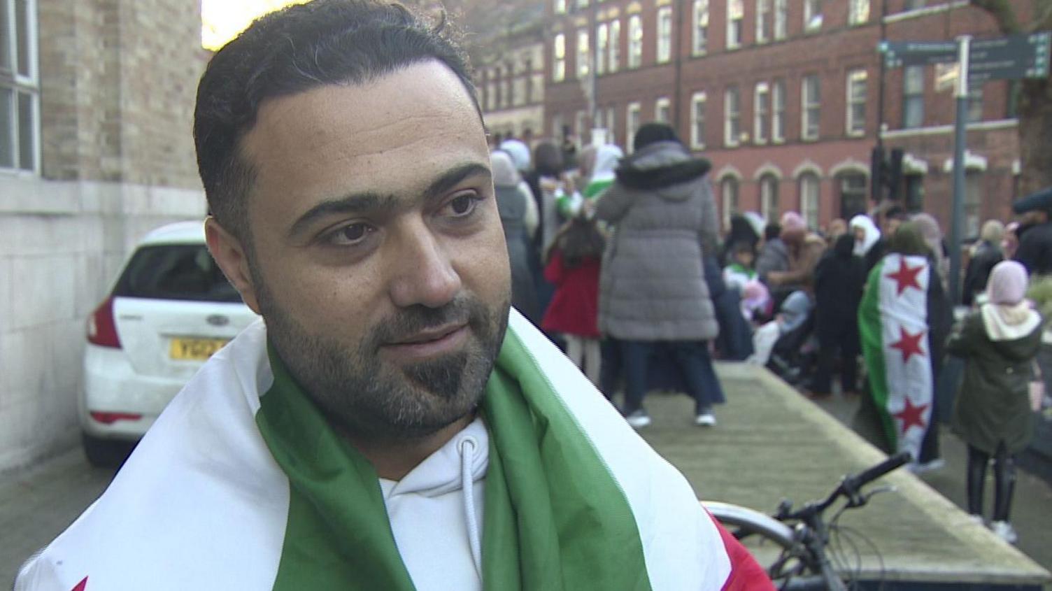 Mazen Haseno draped in a Syrian flag, stood outside broadcasting house with a crowd of people visible in background celebrating
