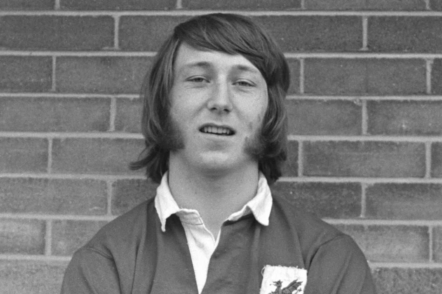 Black and white photo of JPR Williams as a young man, wearing a Wales rugby shirt, standing in front of a brick wall. His hair is shoulder-length and he has large sideburns. 