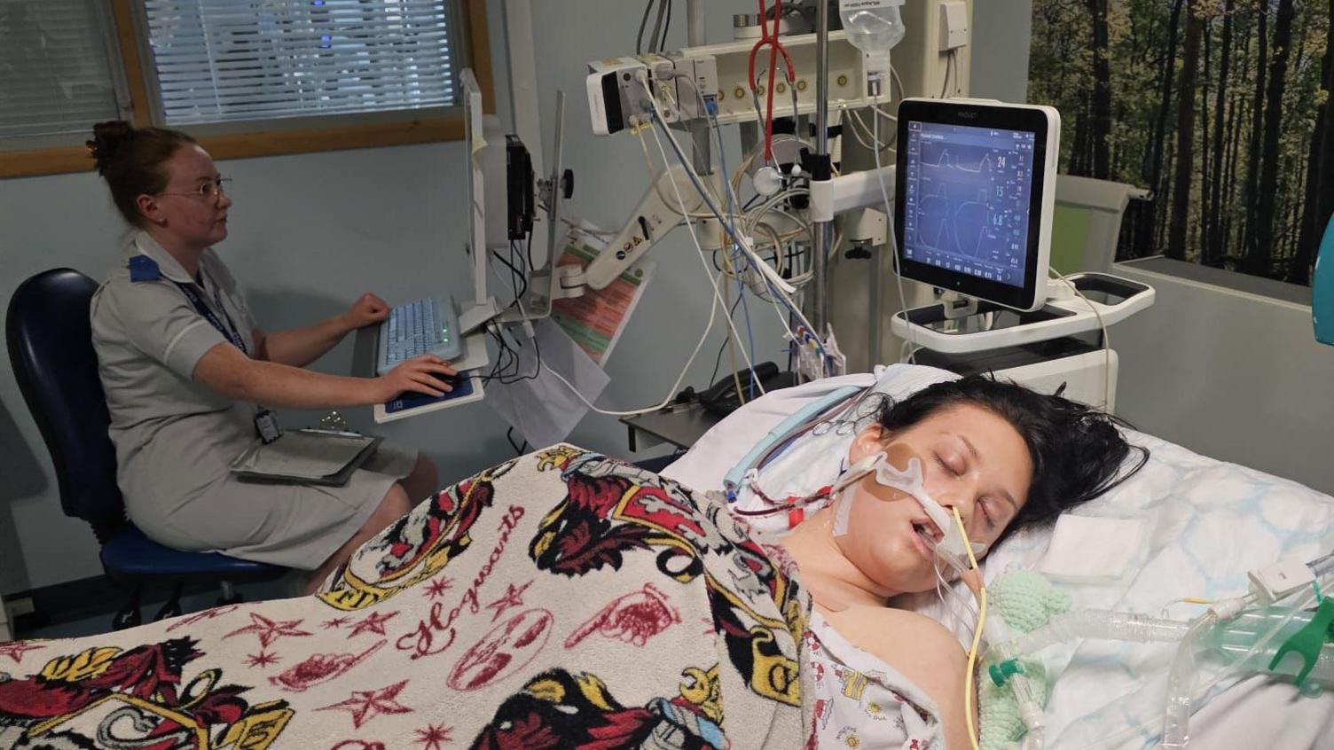 Lilly is show lying in a hospital bed, she has tubes coming out of her mouth and nose and is connected to life support machines. She has a Harry Potter blanket pulled over her with the word 'Hogwarts' on it. A nurse is sat on a chair next to her using a computer. 