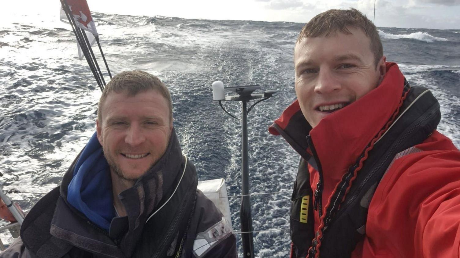 Llun o Thomas a Nick ar y cwch ar y mor. Mae'r ddau yn gwisgo cot a siaced achub 
