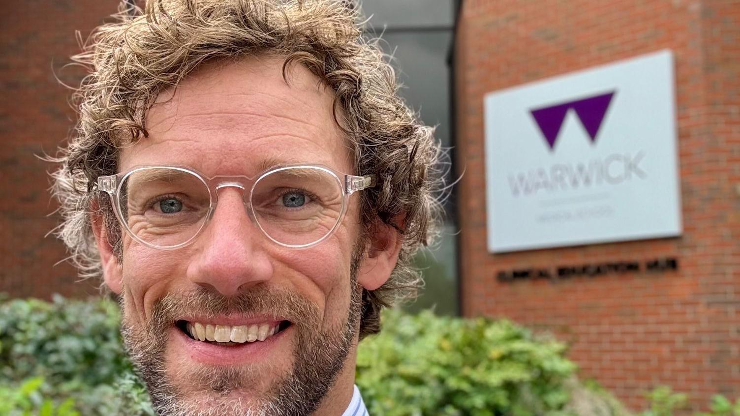 James Gill, a man with a beard, wearing glasses and a blue and white striped shirt. He is stood outside a red brick building with The University of Warwick logo on it.